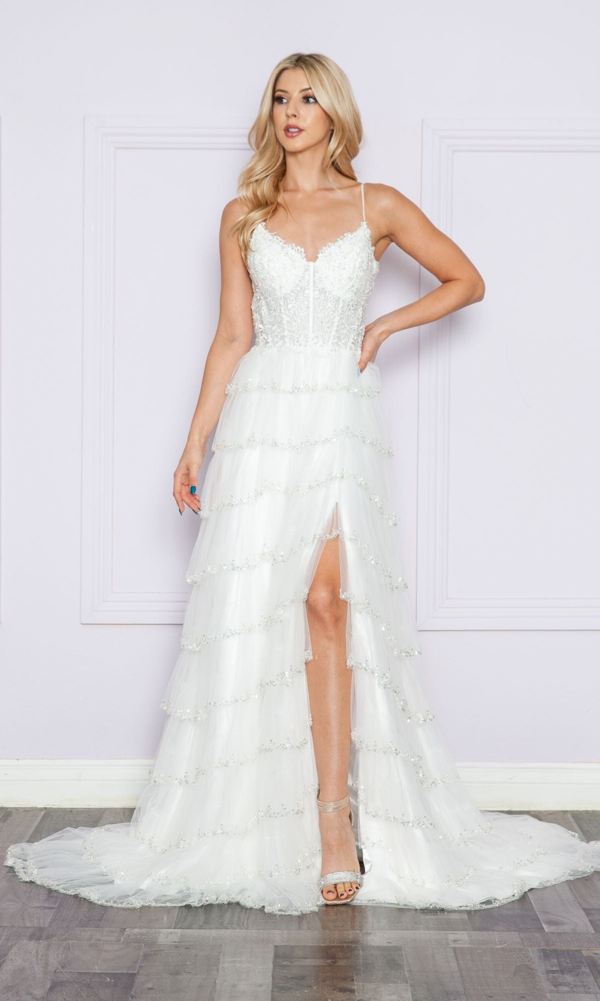A woman stands against a light purple wall wearing the Ruffled Long A-Line Tiered Prom Dress: Poly 9404 with thin straps and a front slit, paired with silver high-heeled sandals.
